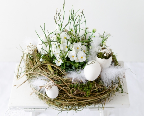 Osternest in Weiß mit Hornveilchen bepflnazt und mit Gänseeiern und Federn dekoriert auf einem alten weißen Holzhocker