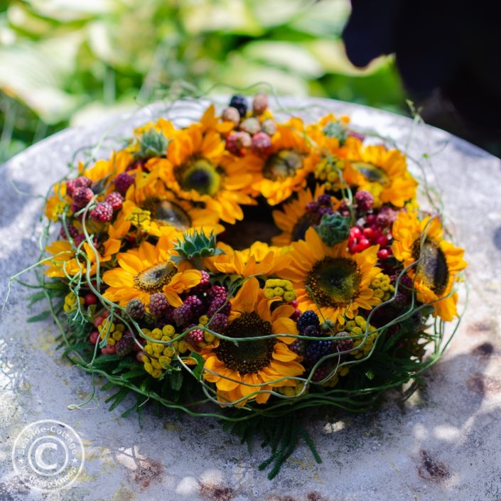 gesteckter-sommerkranz-mit-sonnenblumen-brombeeren-rainfarn-titel Kranz mit Sonnenblumen auf rundem Gartentisch