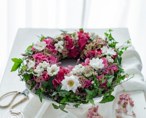 rosa Hortensienkranz mit Herbstanemonen auf Tisch mit weißer Tischdecke vor einem Fenster mit weißer Gardine