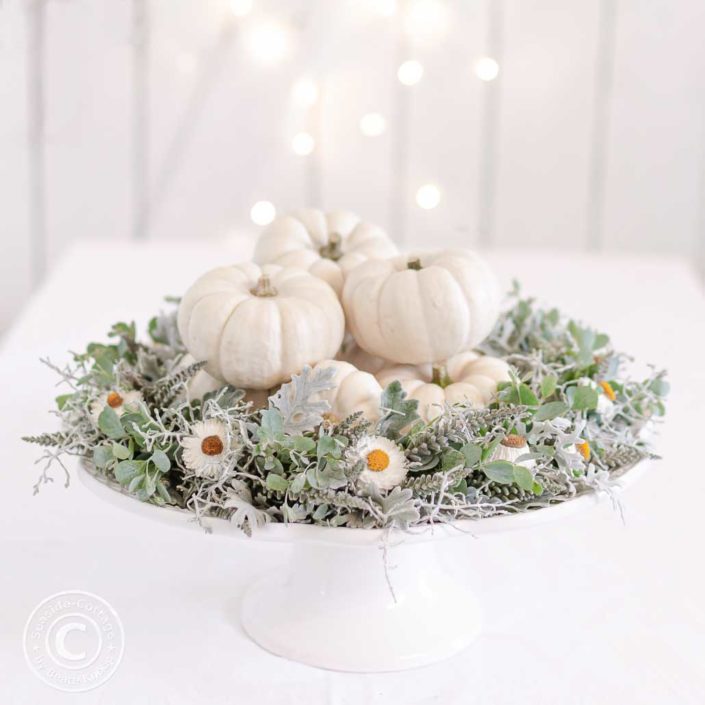 herbstkranz-grau-weiss-mit-baby-boo 03 weiße Tortenplatte auf Fuß dekoriert mit einem grau-weißen Herbstkranz und kleinen weißen Zierkürbissen, im Hintergrund das Bokeh einer Lichterkette vor einer weißen Bretterwand